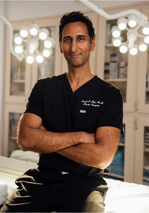 A vertical portrait of Dr. Surjit Rai, a man with dark hair, sitting with his arms crossed on a white examination or operating table. He is wearing black scrubs with "Surjit S. Rai, M.D. Plastic Surgery" embroidered on the chest. He is smiling and looking directly at the camera. Bright, circular operating room lights are visible in the background.