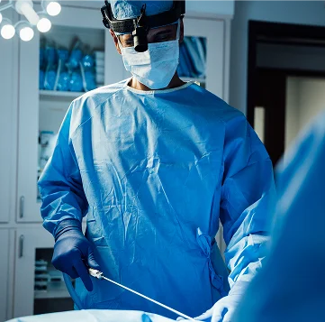 A vertical image of Dr. Surjit Rai, a male surgeon, wearing a blue surgical gown, mask, and a headlamp, performing liposuction. He is holding a cannula, which is inserted into the patient's body in the foreground, while he concentrates intently on the procedure. The operating room is dimly lit with bright overhead surgical lights.