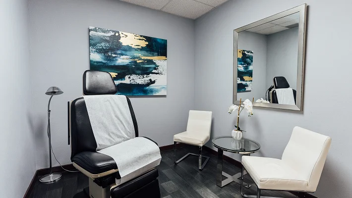 An inside shot of a check up/ procedure room at Cosmetic Surgical Center. Theres a abstract piece of art hangs on the back wall and orchids are seen sitting on the side table between two white chairs.