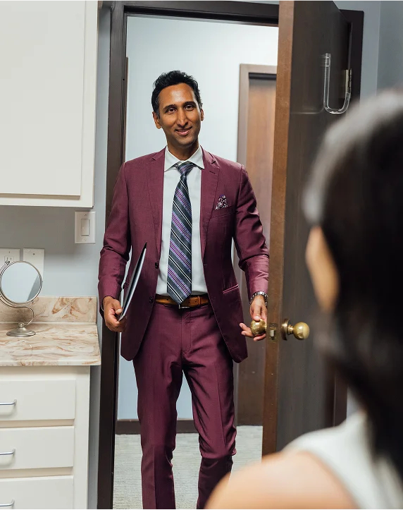 A vertical image of Dr. Surjit Rai, a man with dark hair, wearing a maroon suit and a striped tie, standing in a doorway and smiling. He is holding a dark folder or tablet under one arm and appears to be stepping into a room or holding the door open for a patient. A small vanity area with a mirror is visible on the left, and the shoulder and back of a patient are visible in the foreground.