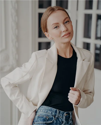 A vertical portrait of a woman with red hair tied back, posing in a bright, elegant interior space. She is looking directly at the camera with a subtle smile and is wearing a cream-colored blazer over a black top and blue jeans. Her right hand is near her collar, and her left hand is resting on her hip. Large, white-framed windows are visible in the soft-focus background.