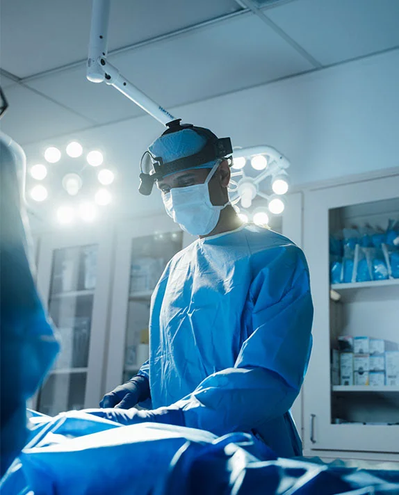 A vertical, dramatically lit image of Dr. Surjit Rai, a male surgeon, standing over an operating table in a surgical room. He is wearing a blue surgical gown, mask, cap, and a headlight. The intense overhead surgical lights illuminate the scene in a cool blue and white tone, focusing on the serious concentration of the doctor. - Sculpting Defined Pectoral Contours in Dallas, TX