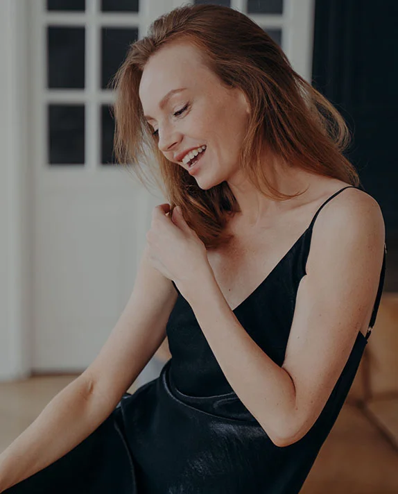 A light-skinned woman with shoulder-length reddish-brown hair is seated, smiling warmly and looking slightly to her left. She wears a dark, likely black, slip dress or top with spaghetti straps. Her joyful expression is complemented by soft, natural lighting. The background shows a light-colored wall and windows, creating a cozy, inviting atmosphere. - Arm Lift in Dallas, TX