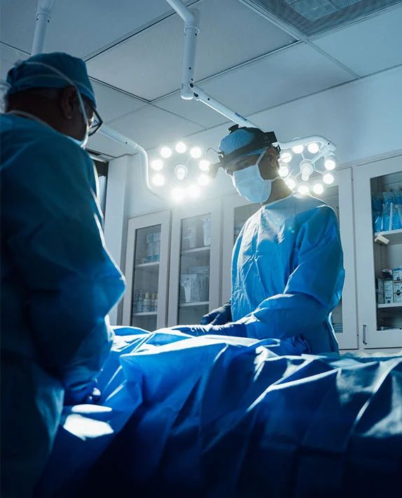 A dark, vertically cropped image showing two surgeons, Dr. Surjit Rai and Dr. Vasdev Rai, performing a procedure in an operating room. Both are wearing blue surgical gowns, caps, and masks. One surgeon, wearing a surgical headlamp, is positioned at the head of the operating table, looking down at the draped patient. The other surgeon is standing on the left side, facing the first surgeon. Bright overhead surgical lights illuminate the scene. - Breast Reduction in Dallas, TX