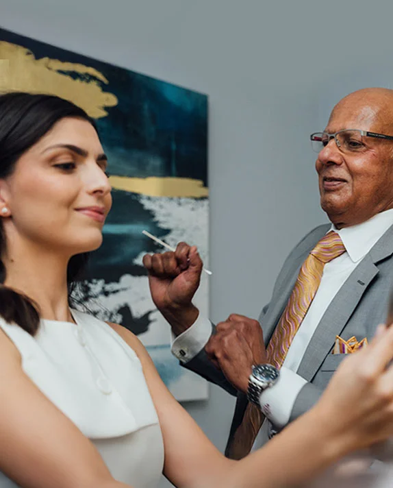 A vertically cropped close-up image showing Dr. Vasdev Rai, an older man with glasses and a bald head wearing a gray suit and a patterned tie, interacting with a female patient. The patient, a younger woman with dark hair and a white top, is smiling and looking off-camera. Dr. Rai appears to be gesturing near her face, possibly during a consultation or examination for cheek implants. A modern abstract painting is visible on the wall in the background. - Cheek Implants in Dallas, TX