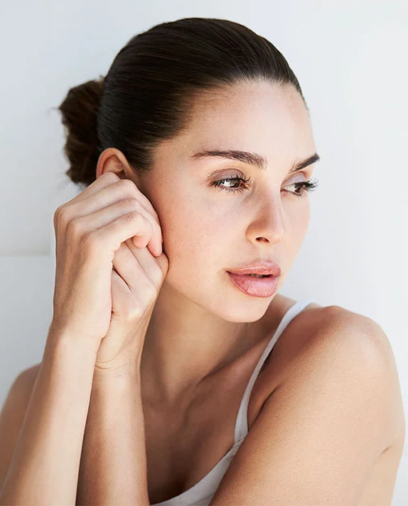 A vertically cropped, close-up image of a woman with dark hair pulled back in a bun, looking away from the camera. She is wearing a white tank top or camisole and is touching the side of her face or ear with both hands. The background is a bright white. - Chemical Peels in Dallas, TX