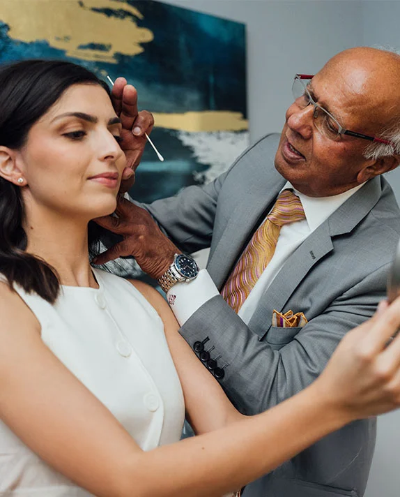 A vertically cropped close-up image showing Dr. Vasdev Rai, an older man with glasses and a bald head wearing a gray suit and a patterned tie, interacting with a female patient. The patient, a younger woman with dark hair and a white top, is smiling and looking off-camera. Dr. Rai appears to be gesturing near her face, possibly during a consultation or examination for cheek implants. A modern abstract painting is visible on the wall in the background. - Eyelid Surgery in Dallas, TX