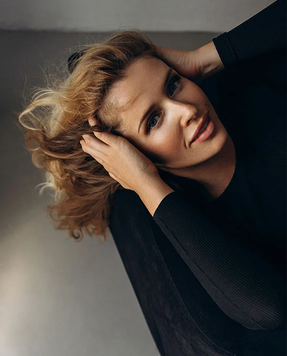 A woman with light brown, tousled hair is viewed from an overhead, slightly angled perspective. She holds her hair with both hands, her head tilted back slightly, and her expression relaxed. She has light skin and light blue eyes. She wears a fitted black top, and the background is a muted gray surface with a soft-focus effect. - Forehead Browlift in Dallas, TX