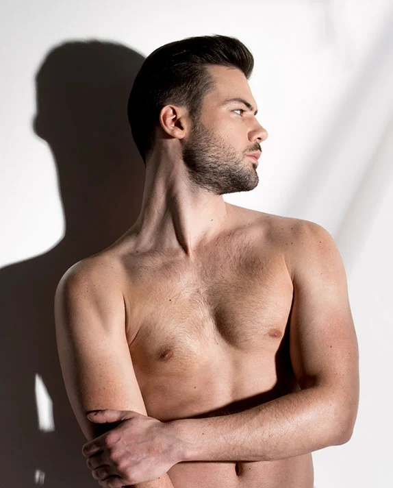 A vertical, shirtless portrait of a man with dark hair and a beard, looking to the right. He is posing against a white wall with his arms crossed over his chest, and dramatic, harsh side lighting creates a prominent shadow of his head and shoulder on the wall behind him. - Gynecomastia in Dallas, TX