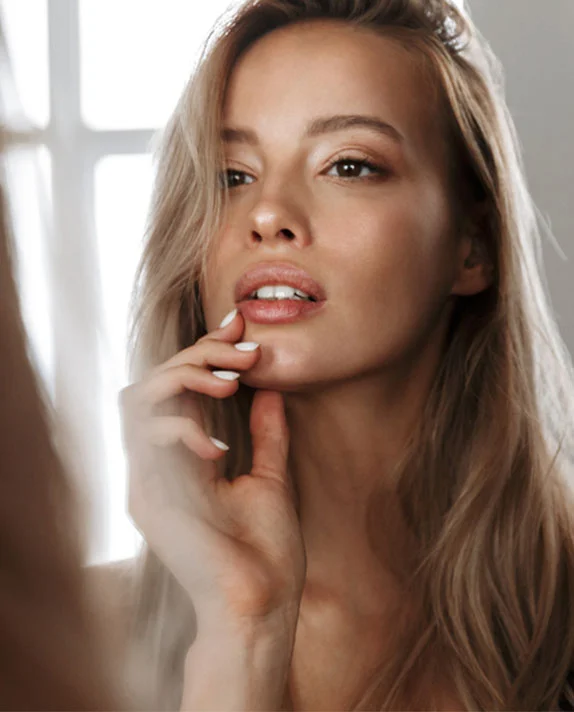 A woman with long, blonde hair looks at her reflection in a mirror, her hand gently touching her chin and jawline. Her lips are slightly parted, and she has light eye makeup and a natural glow. The background is bright and softly blurred, with a hint of a window frame. - Lip Fillers in Dallas, TX