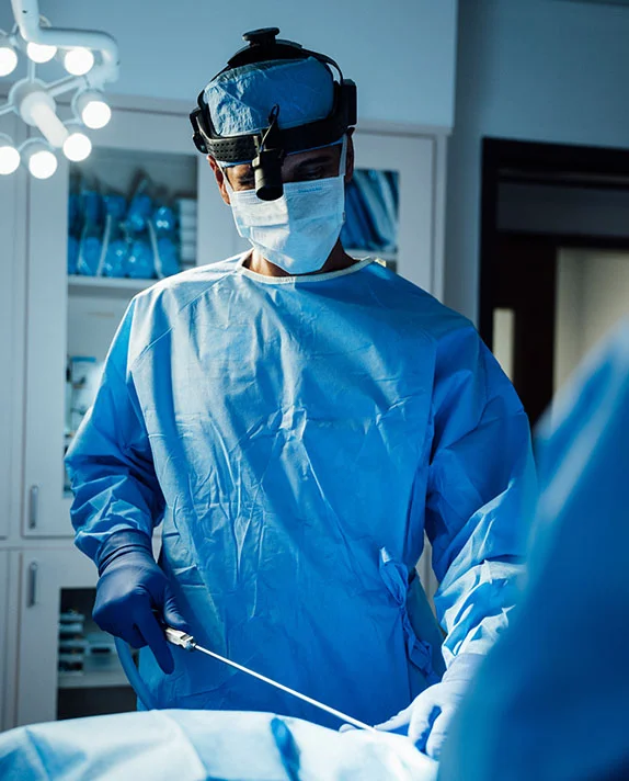A vertical image of Dr. Surjit Rai, a male surgeon, wearing a blue surgical gown, mask, and a headlamp, performing liposuction. He is holding a cannula, which is inserted into the patient's body in the foreground, while he concentrates intently on the procedure. The operating room is dimly lit with bright overhead surgical lights. - Liposuction in Dallas, TX