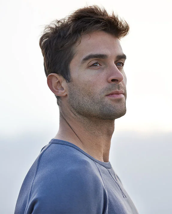 A bright, vertical outdoor portrait of a handsome young man with dark, slightly messy hair and light stubble, wearing a blue-gray crewneck shirt. He is looking up and to the side with a pensive expression against a soft, bright, and slightly blurred background. - Male Botox in Dallas, TX