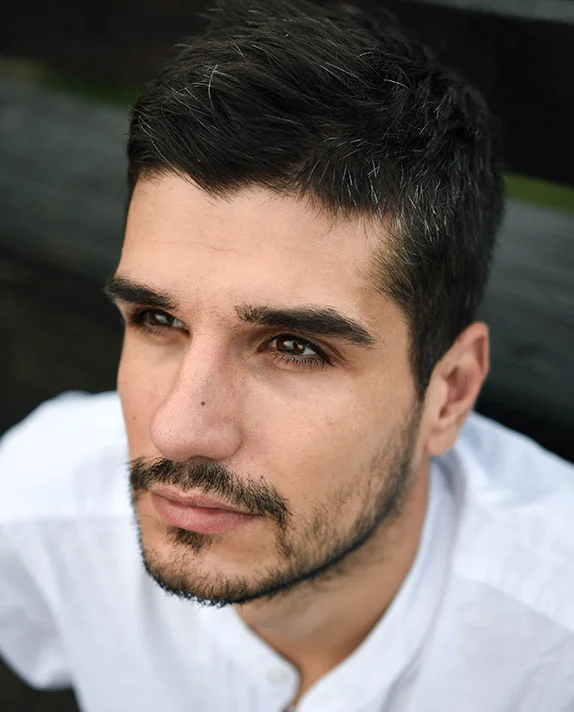 A close-up vertical portrait of a handsome young man with dark hair, a stubble beard, and expressive brown eyes, looking upward and to the side with a serious expression. He is wearing a white collared shirt, and the background is dark and out of focus. - Male Browlift in Dallas, TX