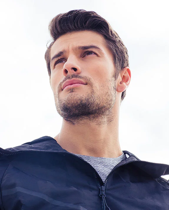 A low-angle, vertical portrait of a handsome young man with dark, styled hair and stubble, looking up with a determined expression. He is wearing a black zippered jacket over a gray shirt, set against a bright, featureless sky. - Male Chin Augmentation in Dallas, TX