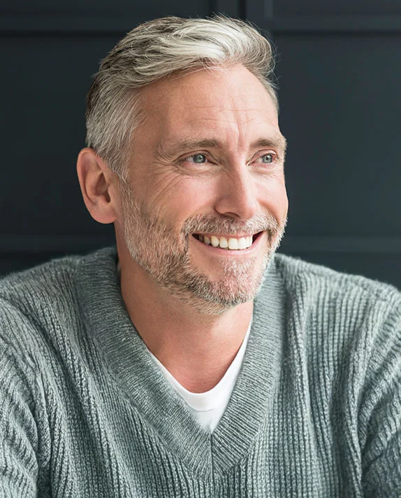 A cheerful vertical portrait of an attractive middle-aged man with gray hair and a short beard, smiling broadly as he looks off to the side. He is wearing a cozy gray V-neck sweater over a white shirt, set against a dark, featureless background. - Male Eyelid Surgery in Dallas, TX