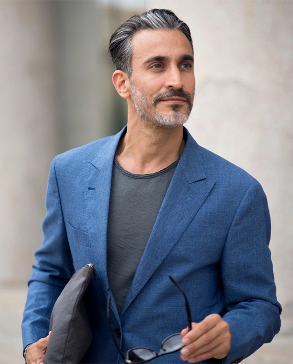 A vertical portrait of an attractive middle-aged man with dark, graying hair slicked back. He is wearing a bright blue sport coat over a dark gray T-shirt and holding sunglasses in one hand, looking off to the side. - Male Facelift in Dallas, TX