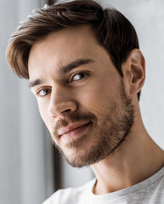 A close-up of a woman with short dark hair, wearing a white shirt and small gold hoop earrings. She is resting her chin on her hand and looking at the camera. The background is a solid white. - Male Otoplasty in Dallas, TX