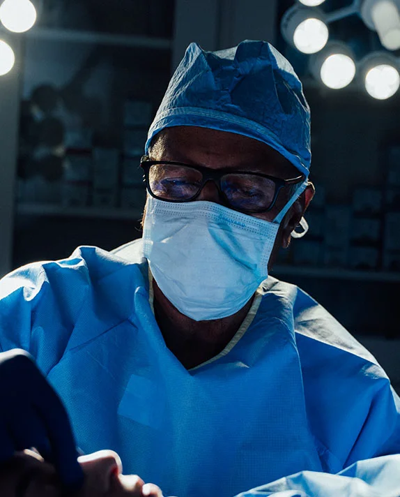 A close-up, vertical image of Dr. Vasdev Rai, an experienced surgeon, in an operating room setting. He is wearing a blue surgical gown, cap, face mask, and glasses, concentrating intently on a procedure, with bright, circular surgical lights providing dramatic, cool illumination in the dark background. - Male Rhinoplasty in Dallas, TX