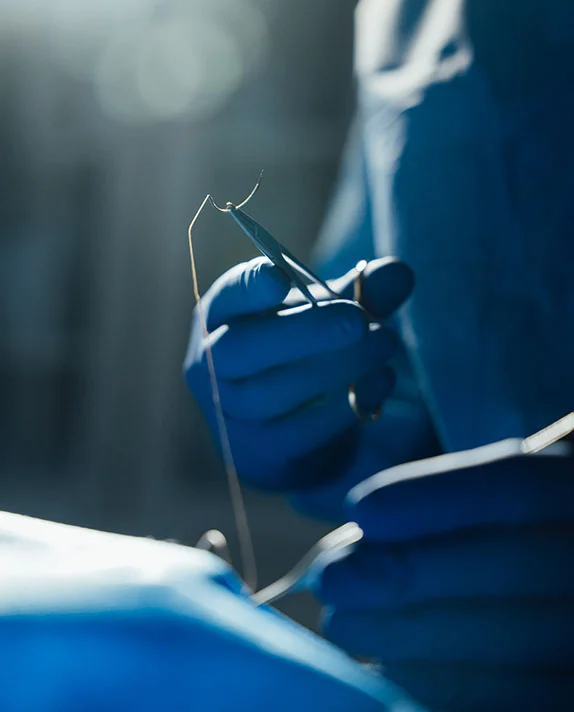 A vertical close-up image showing a surgeon's hands, wearing bright blue surgical gloves, performing a procedure with a needle and thread held by forceps. The scene is darkly lit with a strong focus on the instruments and the gloved hands, highlighting the detail of the surgical work, likely for a tummy tuck or other body contouring procedure. - Male Tummy Tuck in Dallas, TX