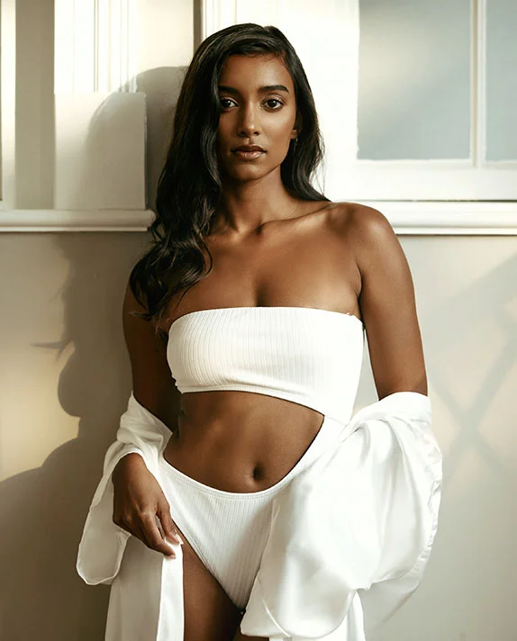 A young woman with long, dark hair, wearing a white strapless top. She is looking directly at the camera with a neutral expression on her face, and sunlight is shining from the left on her face and shoulder. The background is a white wall with a vertical molding. - Mommy Makeover in Dallas, TX