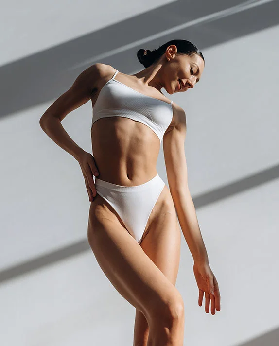 A woman is shown from the mid-section up in a three-quarter view, lit from above and the side. The light casts long, thin striped shadows across the light gray wall behind her. She wears a simple white lingerie set consisting of a supportive crop top and fitted briefs. Her posture is relaxed yet confident, with arms slightly bent and one hand resting near her waist. Her gaze is directed downward, expression soft, hair pulled back in a bun. Her smooth, even-toned skin and toned form are highlighted by the lighting. - Thigh Lift in Dallas, TX