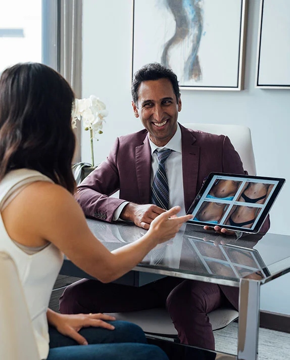 A vertical image of Dr. Surjit Rai, a male doctor wearing a maroon suit and tie, sitting behind a glass desk and smiling warmly at a female patient. He is showing her four photos of abdomens, likely tummy tuck before-and-after results, on a tablet screen, while the patient points to the screen. The office setting is modern, with abstract art on the wall behind him. - Tummy Tuck in Dallas, TX