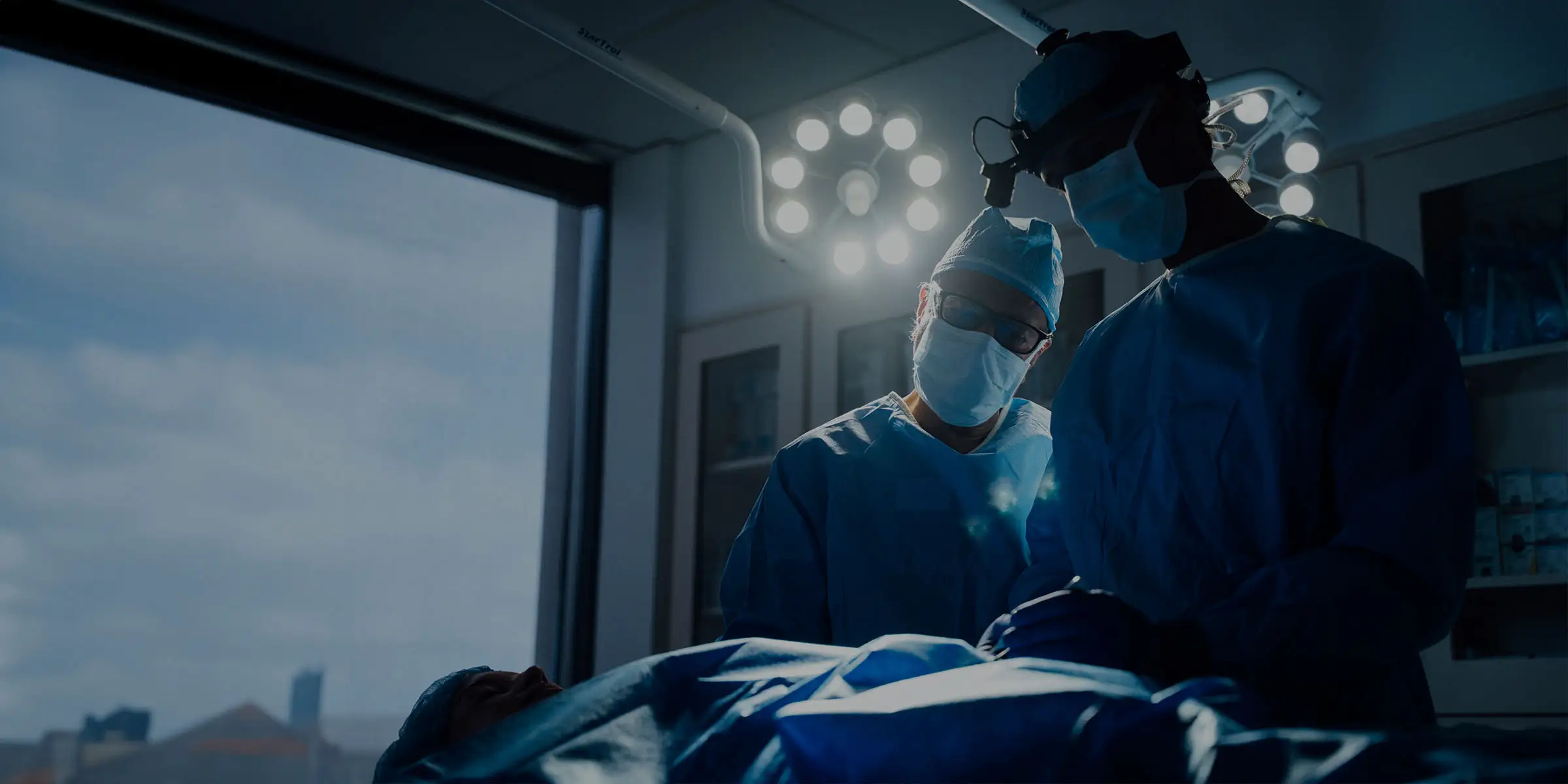 A cinematic horizontal banner image showing two surgeons, Dr. Surjit Rai and Dr. Vasdev Rai, performing surgery on a patient, likely rhinoplasty, in a dimly lit operating room. Both doctors are wearing blue surgical gowns, masks, and caps, and are illuminated by the intense overhead surgical lights. One surgeon, wearing glasses, is leaning over the patient's face, while the other stands slightly behind him.
