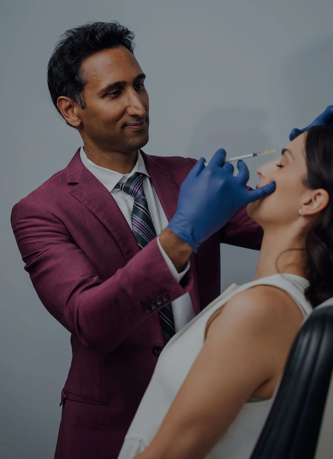 A vertical image of Dr. Surjit Rai, a male doctor wearing a maroon suit and tie with blue surgical gloves, administering an injection, likely a liquid facelift or dermal filler, to a female patient. The patient is lying back in a treatment chair with her eyes closed as the doctor works near her eyebrow, with a light gray background.
